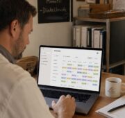 Ein Bäckereiinhaber sitzt an einem Schreibtisch und erstellt am Laptop einen digitalen Schichtplan. Daneben stehen eine Kaffeetasse, Notizen und ein Smartphone, im Hintergrund ist die Backstube zu sehen.