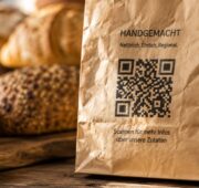 Nahaufnahme einer Bäckertüte aus braunem Papier mit aufgedrucktem QR-Code. Im Hintergrund sind unscharf verschiedene Brote und Brötchen in einer warm beleuchteten Bäckerei zu sehen. Auf der Tüte befindet sich ein Hinweis zum Abrufen von Zutateninformationen per QR-Code.