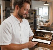 Ein Bäcker in weißer Arbeitskleidung steht in einer Backstube und betrachtet zufrieden ein Tablet mit einer Bäckerei-Website. Im Hintergrund sind Backöfen, Arbeitsflächen und frisch gebackene Brote zu sehen.